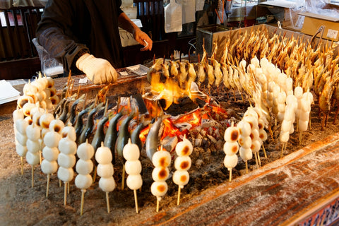 Around a charcoal fire, a stall vendor roasting skewered Iwana fish and Kushi Dango