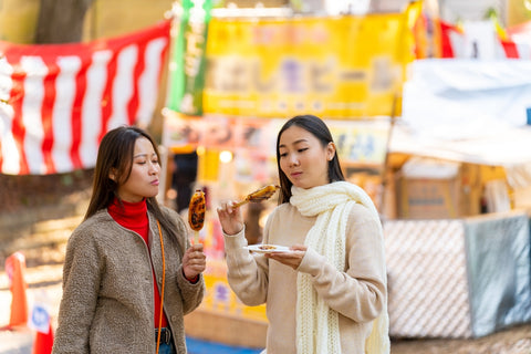 Asian woman friends eating grilled mochi with soy sauce on sticks together while travel at at street market in Tokyo city, Japan.