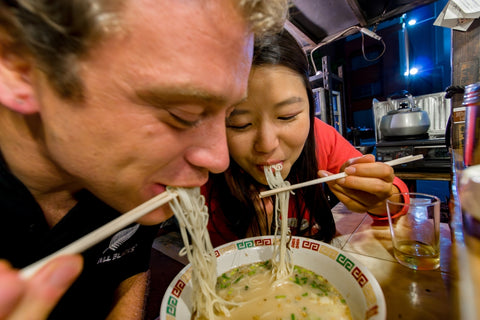 Yatai food stalls, Naka river bank, Western tourist and asian friend eating together a bowl of Hakata Ramen (also called Tonkotsu), famous local food speciality as a Japanese Street Food