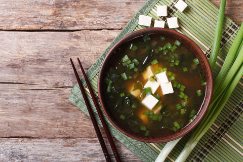 A bowl of miso soup is surrounded by tofu chunks, green onions, and a pair of chopsticks atop a green placemat.