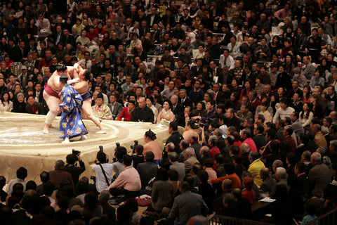Two sumo wrestlers engaging while photographers take pictures and the audience cheers in the Tokyo Grand Sumo Tournament