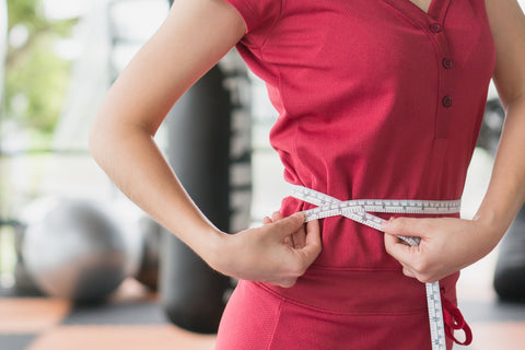 Young slim woman measuring her waist by measure tape after a diet with accessory in sporty gym as background.