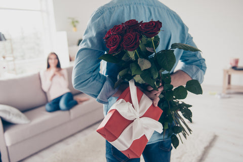Unexpected moment celebrated in routine everyday life! Cropped photo of man's hands hiding holding chic bouquet of red roses and gift with white ribbon behind back, happy woman is on blurred background