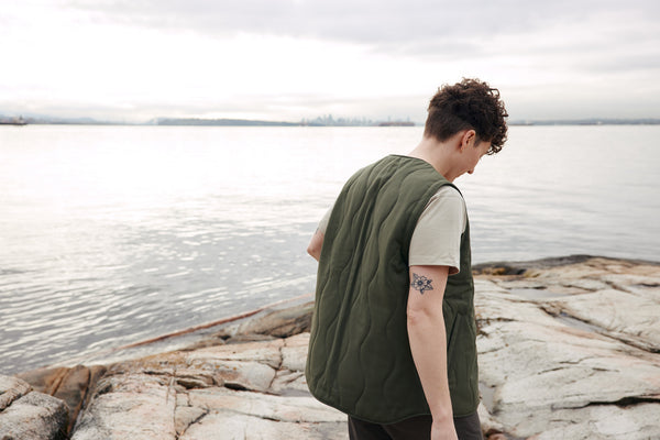 AK MacKellar wears the ecologyst Quilted Wool Vest in Moss Green and Heavyweight Tee in Oyster with the ocean and Vancouver skyline in the background.