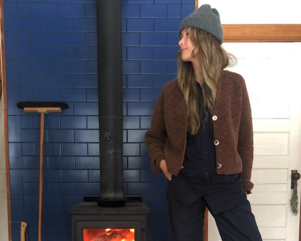 Leah Gibson wears the Port Cardigan in Speckled Sunset as she stands in front of a wood burning fire