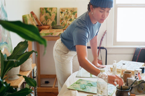 Robyn Jin paints in her home studio wearing the Merino Tee in Blue, the Fisherman Toque in Speckled Blue, and the Women's Canvas Work Pant in Salt