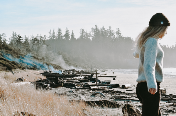A woman wearing a Fisherman Sweater walks across a driftwood log in search of a wave.