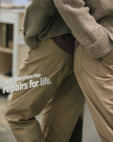 A person wearing our Fisherman Full Zip in Speckled Cream and the Work Pant in Hazelnut leans against a mirror that says, "hey! that piece has repairs for life."