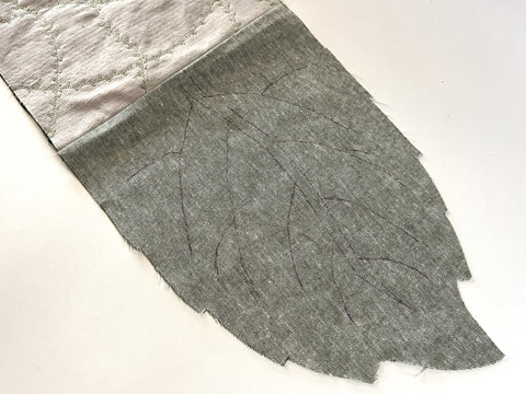 The assembled pieces are shown - a light-green leaf shaped piece with quilting lines marked, sewn to the tan fabric. It is ironed flat.