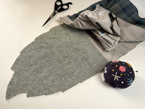 An in-process photo of attaching the Satchel Front to the assembled Flap + Back is shown. There is a pair of scissors and flowered pincushion sitting on either side of it.