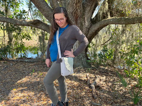 A woman in a blue tshirt, green pants, and black sweater wears a Leaf Satchel across the body. The body of the Satchel is beige, while the flap is light green. Embroidery is visible on the strap.