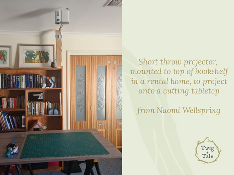 A projector setup by Naomi Wellspring - a short throw projector is mounted to the top of a bookshelf by means of a wooden plank and extended mount arm, in order to avoid holes in the wall. The projector is set up to project onto a cutting table below.