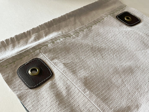 The Satchel Front is shown laid flat but from an angle, focused on where a bronze grommet has been installed through a whole in the leather patch + satchel side. You can see the other patch in the background with a grommet installed as well.