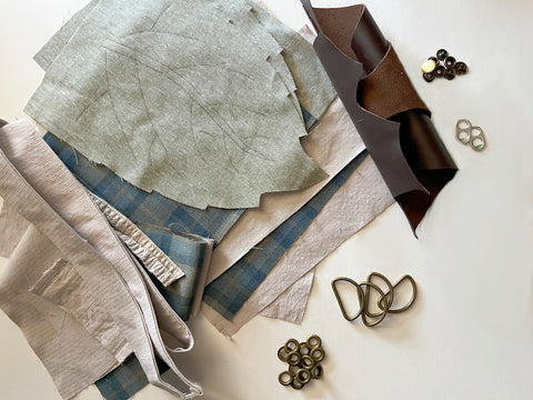 The required materials for the Convertible Hip Pouch hack are shown laid out together, including the cut satchel pieces in tan cotton ripstop material; a light green marled cotton leaf-shaped flap; a blue and green plaid lining upcycled from a suit coat; dark reddish-brown leather; 2 pop tabs; and bronze D-rings, grommets, and snaps.