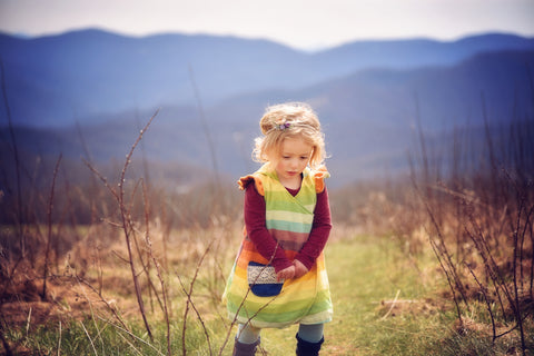 An image of the free Petal Wrap pattern on a young child with blond curls, sewn in rainbow fabric. The child is against a backdrop of mountains and meadow.