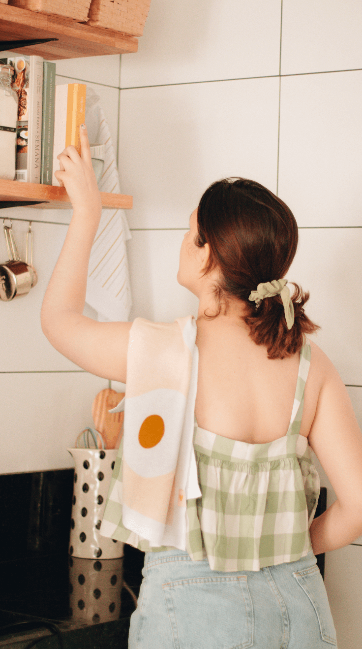 esta imagem mostra a modelo com um pano de prato ovos no ombro. ela está pegando o caderno de receitas meg e meg de uma prateleira e ao fundo podemos ver o pano de prato listras