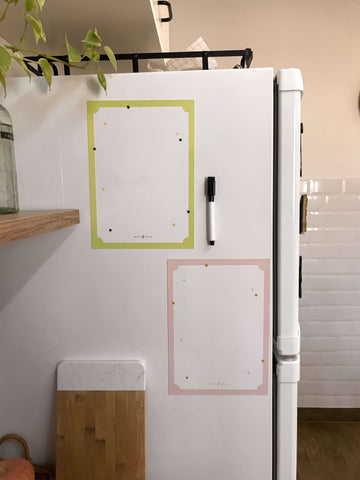 esta imagem mostra uma geladeira com duas lousas magnéticas grudadas nela. também aparecem na imagem uma tábua de madeira e uma planta