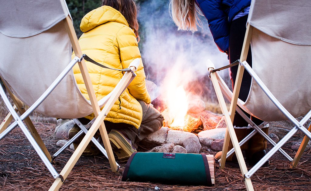 Two people in front of a stone circle campfire; Tom's Firewood Carrier is between them holding some logs.