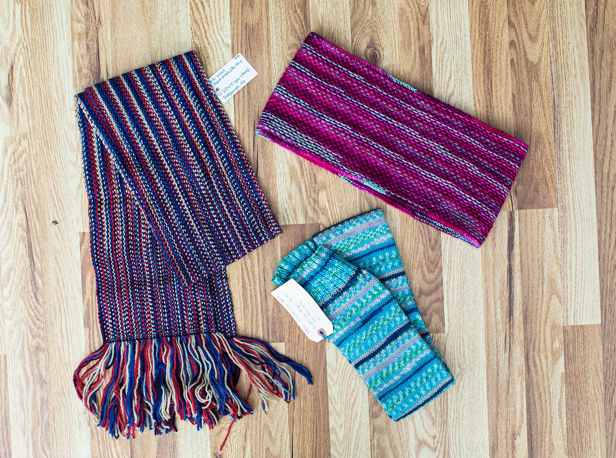 Three crafted items, clockwise from upper right: a vibrant Magenta cowl, a stripy blue scarf, and a scarf in blue/red/green/purple.