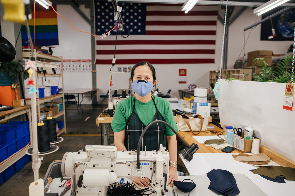 Honguyen, Sewing Supervisor In Training, stands in front of Rainbow, American, and Earth flags at the TOM BIHN factory in Seattle.