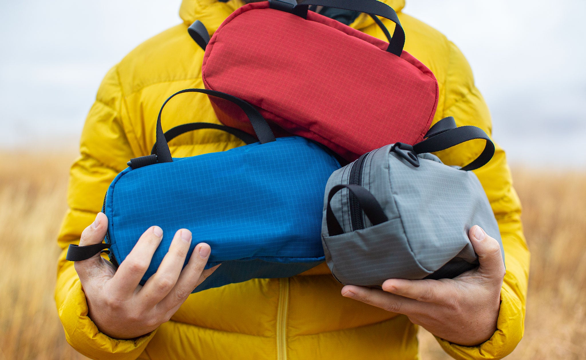 Person wearing a bright yellow down jacket holding three Grab Bags in Cerylon fabric: clockwise, it's Sangria red, Mountain grey, and Cobalt blue.