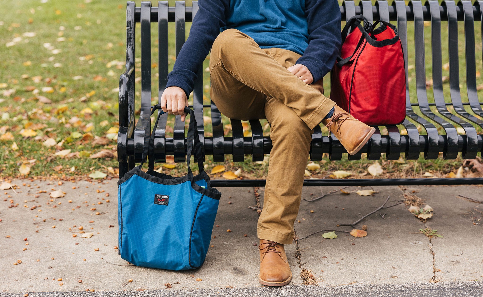 Person sitting on a park bench with Cerylon Original Shop Bags in Cobalt and Sangria.