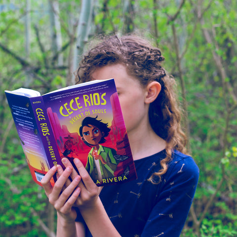 Photo shows girl reading OwlCrate Jr copy of Cece Rios and the Desert of Souls. Girl is standing in green forest.
