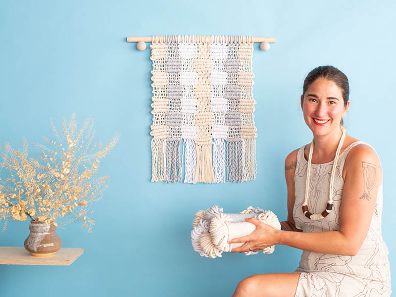Emily Katz and the Hygge Quilt macramé wall hanging, styled on a blue wall with flowers. 