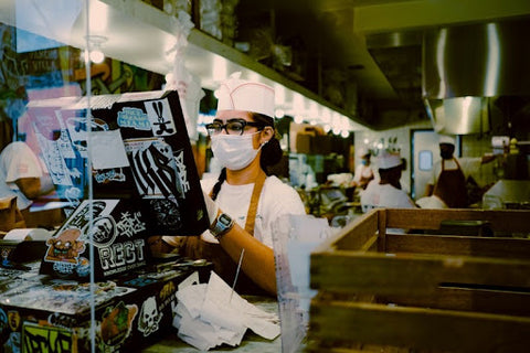 A food service cashier wearing a mask and working behind a clear antimicrobial barrier