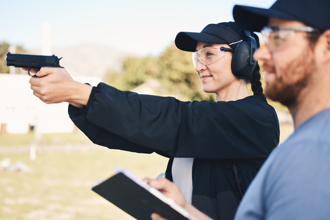 woman with safety equipment aiming gun with trainer's help