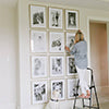 Woman hanging framed photos in wall to ceiling grid gallery style.