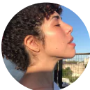 A woman with curly hair basks in the sunlight, eyes closed, background of a clear sky.