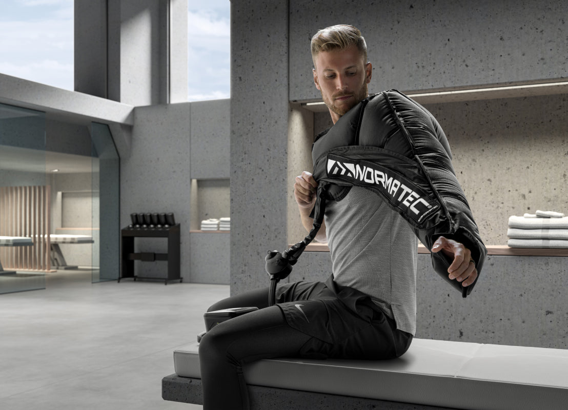 a man on a gray bench wearing workout clothing wearing a Normatec on his left arm