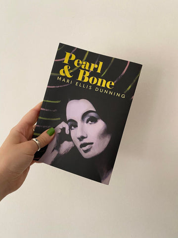 A hand with light green nail polish is holding up a copy of Pearl and Bone which has a painted image of Christine Keeler on its cover.