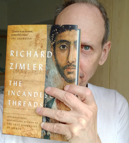 Author Richard Zimler poses with his book The Incandescent Threads, the book is half covering is face making an illusion between his face and the image on the cover