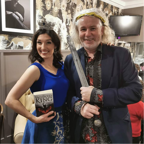 Author Jodie Bon is wearing a blue dress and holding a copy of The Vagabond King next to publishing director Richard Davies who is wearing a navy suit and holding a sword.