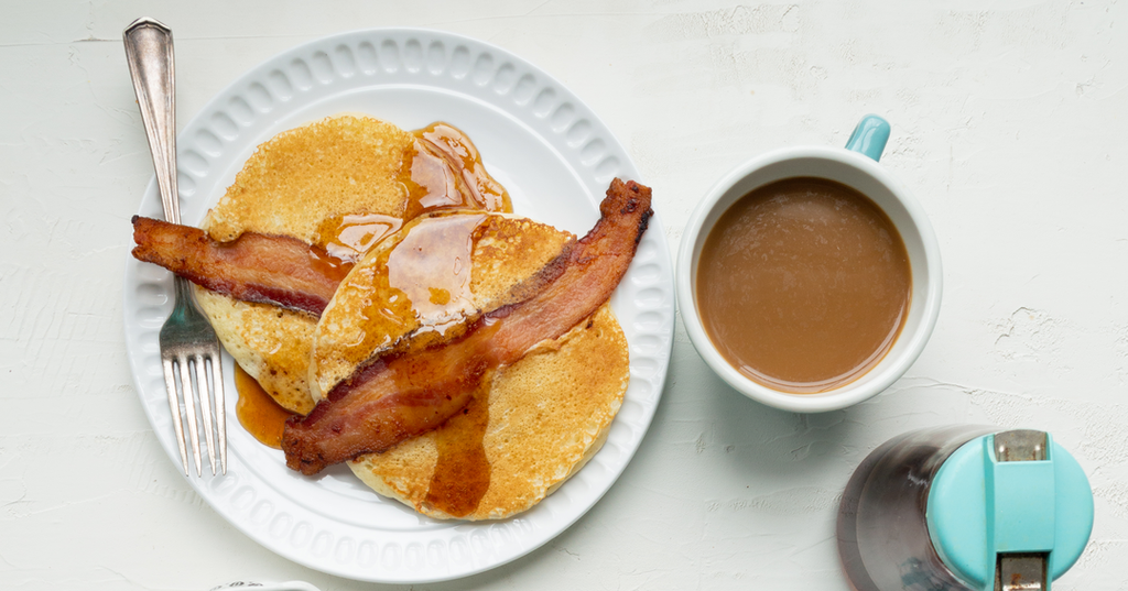 A plate of two overlapping pancakes with a strip of bacon on top of each. A cup of coffee in a white and teal mug is sitting next to the plate.