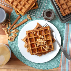 pumpkin flavored waffles sitting on a plate garnished with walnuts surrounded by staged items like cinnamon sticks, syrup, and a Whiskware Batter Mixer