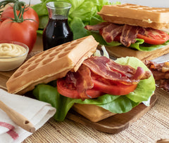 A bacon, Lettuce, and Tomato sandwich between two waffles with vegetables staged in the background with a bottle of syrup