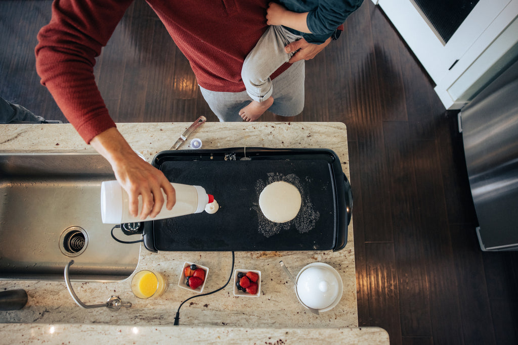 A man holding a toddler with one arm while pouring pancakes batter onto a griddle with the other. We see ramekins of strawberries and maple syrup in front of the griddle. 