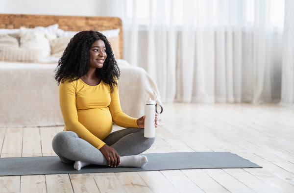 A pregnant woman sits cross legged on a yoga mat, holding a water bottle