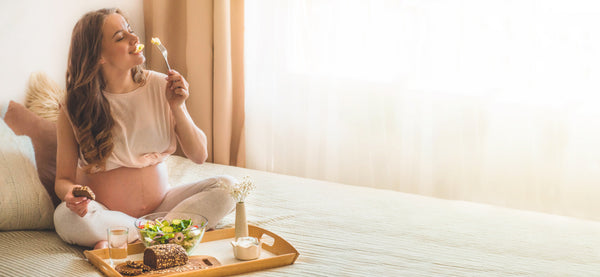 Pregnant woman sitting on a bed, eating a salad and slices of dark bread