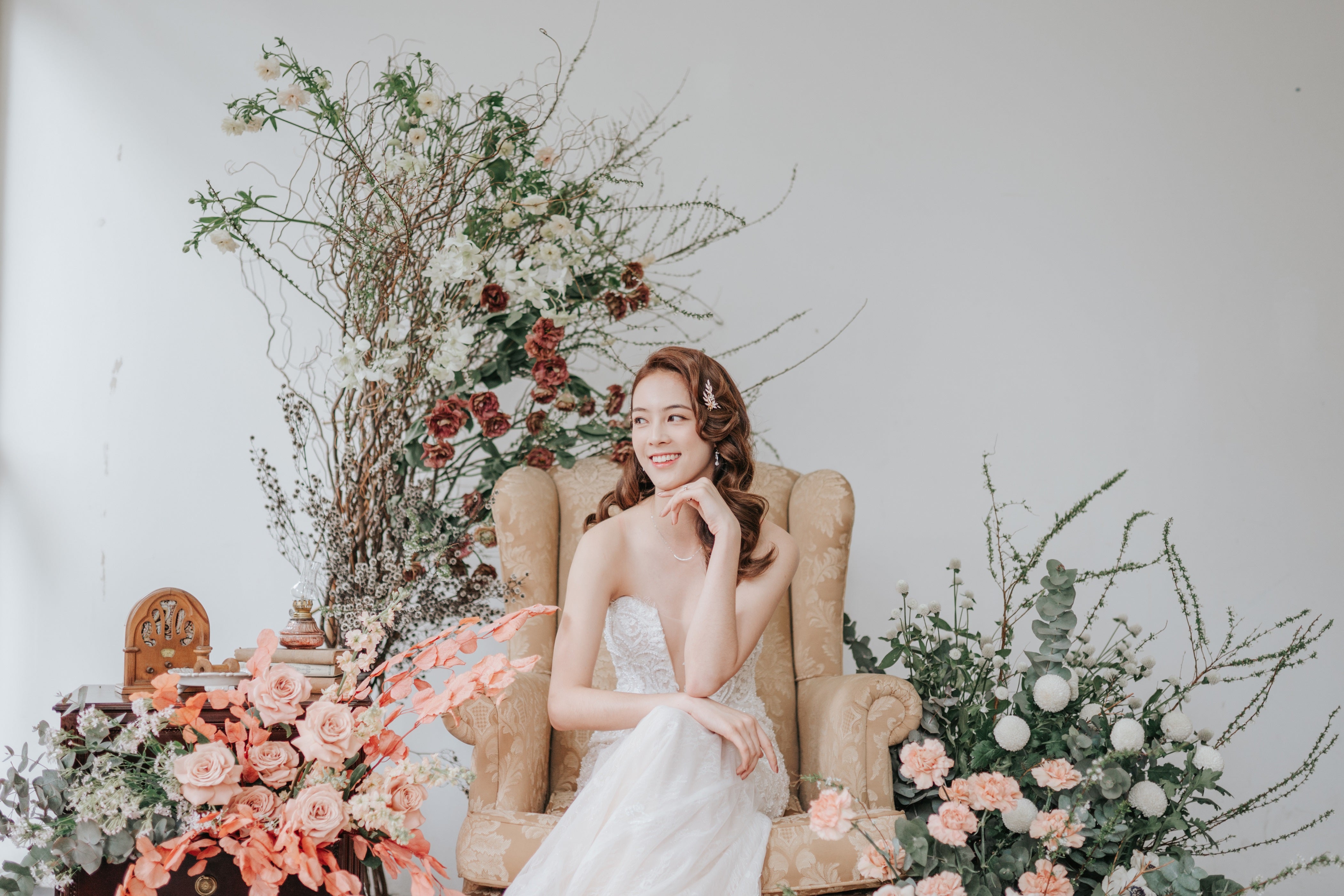 Bride looking sideways, smiling, seated on gold armchair surrounded by arranged flower installation