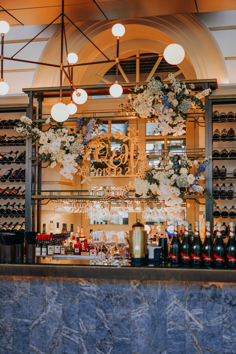 Warmly lit marble bar counter decorated with white orchids, blue hydrangeas, eucalyptus next to wine bottles and glasses at ACM Empress restaurant