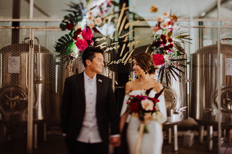 Bride and groom in casualwear standing in front of the brewing vats at Little Island Brewery, bouquet in bride's hand