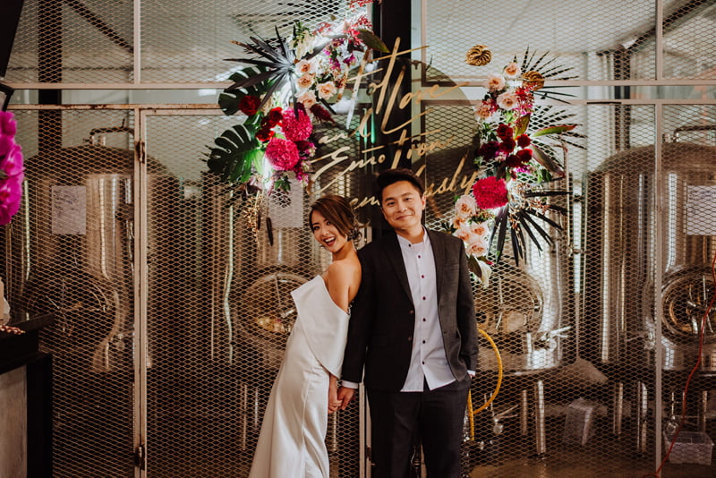 Wedding couple styled in front of dark colour orchids, tropical leaves and fronds, roses arranged in front of beer brewing vats at Little Island Brewery Changi Village