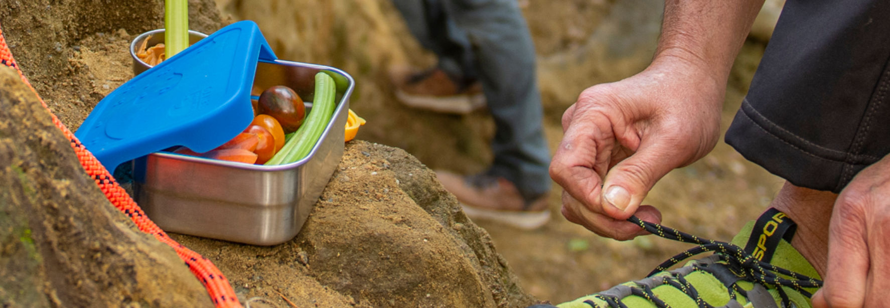 Blue Water Bento container filled with fruits and vegetables on a rock next to someone re-tying their hiking shoe.