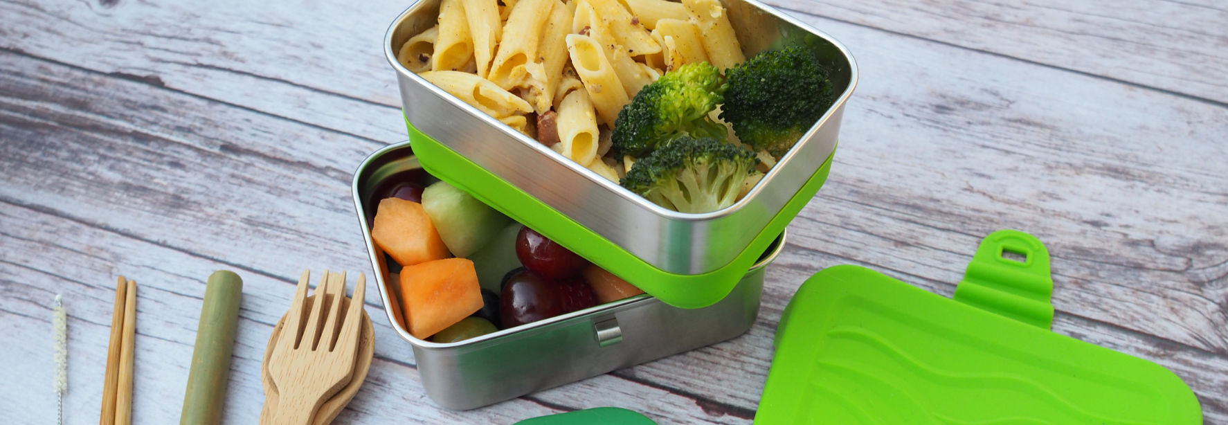 ECOlunchbox Blue Water Bento containers holding pasta, broccoli, and fruits on a table with reusable forks and straws.