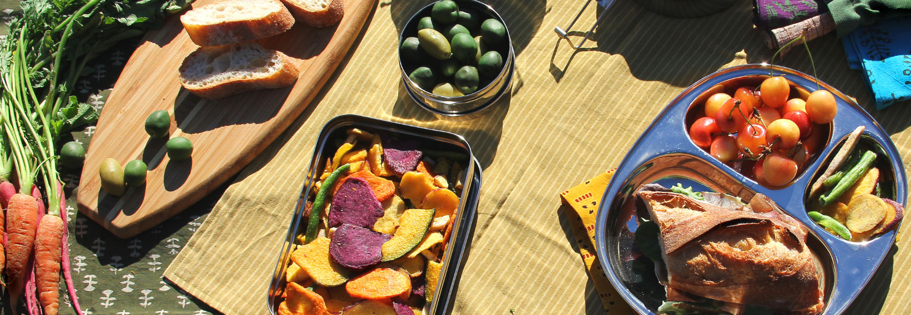 Stainless steel containers and wood serving board with fruits, veggies, and a sandwich laying on colorful picnic blankets.