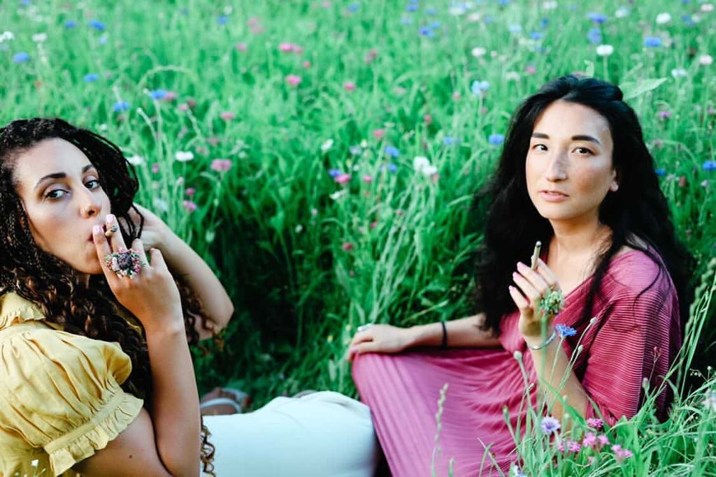 Two women smoking Brown Bear Herbs, organic herbal smoking blends in a field of flowers.